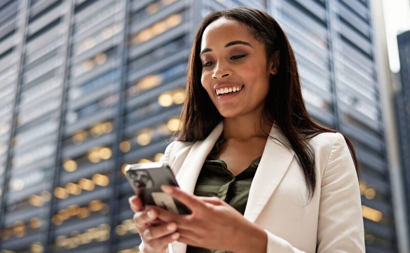 A woman in business professional dress stands outside in front of a skyscraper, using her smart phone. The ubiquity of smart phones is a contributing factor to how social media changed public relations.