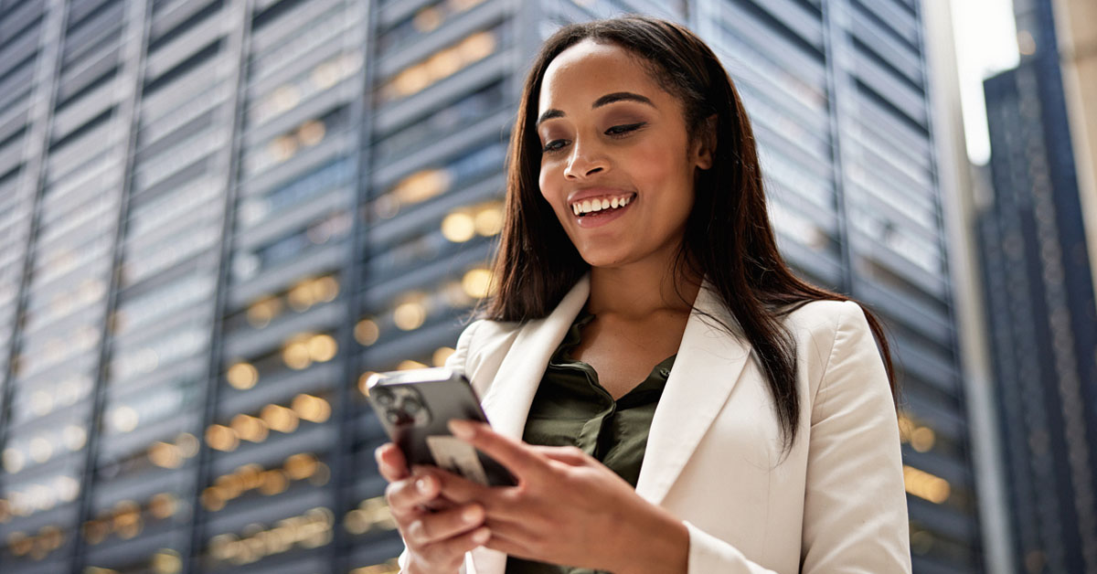 A woman in business professional dress stands outside in front of a skyscraper, using her smart phone. The ubiquity of smart phones is a contributing factor to how social media changed public relations.